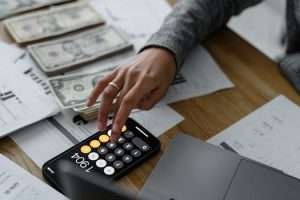 Hand of a Person Using a Calculator Near Cash Money on Wooden Table