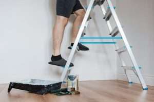 Side view of crop unrecognizable painter in slippers standing on stepladder while renovating room with brush in soft daylight