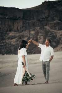 A couple enjoys a romantic moment on a sandy beach with a scenic cliff backdrop.