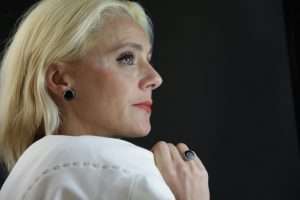 Close-up portrait of a blonde woman with earrings and ring against a dark background.