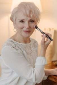 Charming senior woman applying blush with a makeup brush, smiling softly.