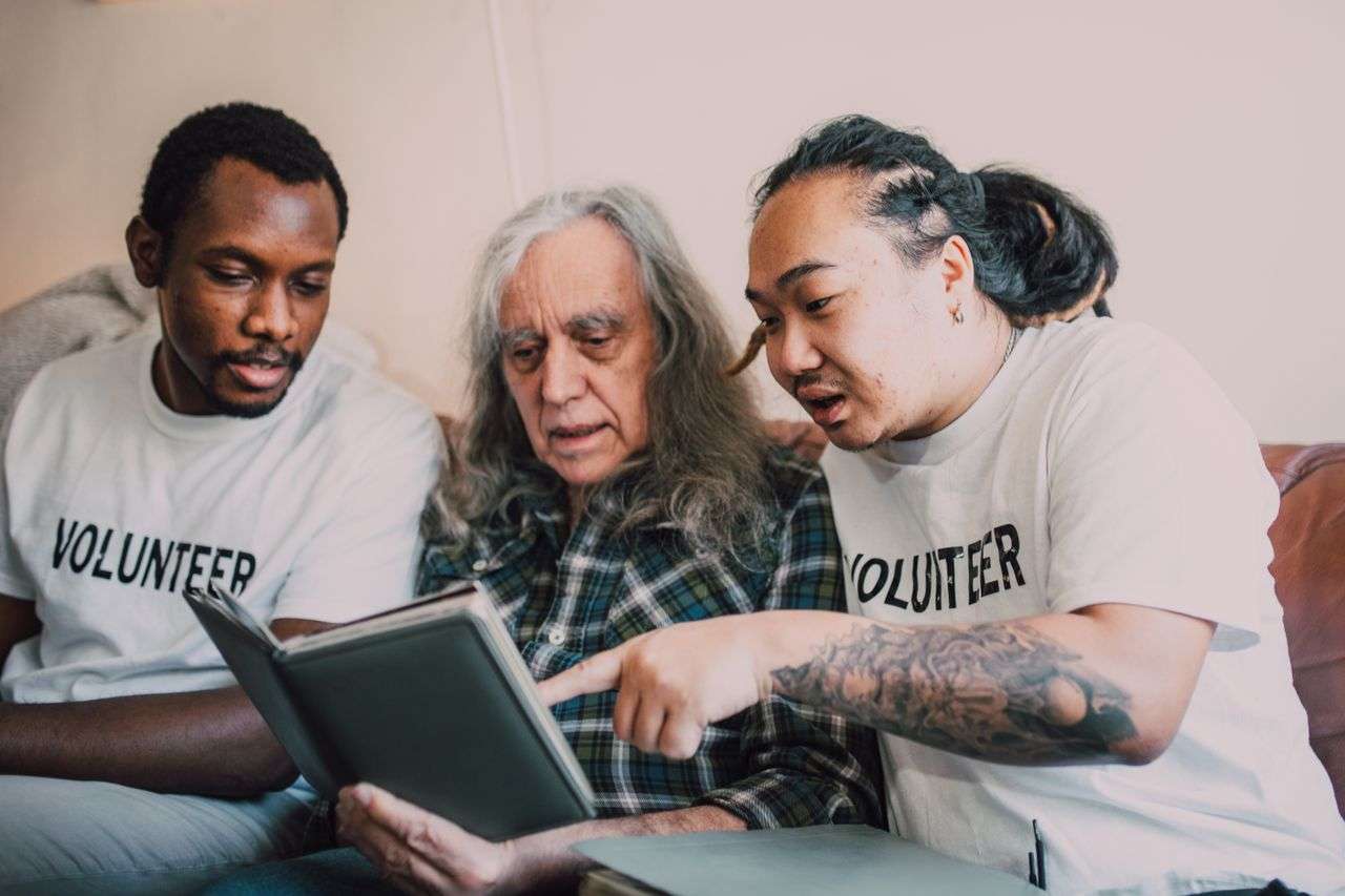 Volunteers assisting an elderly man with a photo album, demonstrating community support and multiculturalism.